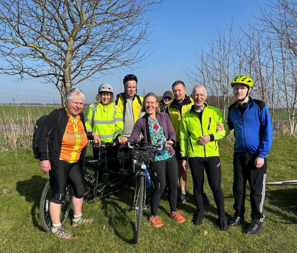 A pre ride group shot of 8 lycra clad cyclists  smiling straight into the camera with grass and trees against a bright blue cloudless sky, in the background