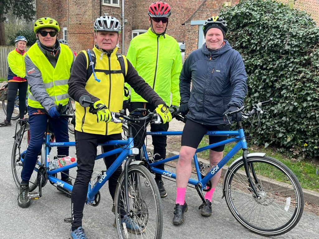 Two tandems side by side with riders aside the bikes in their bright hi-vis jackets and helmets.