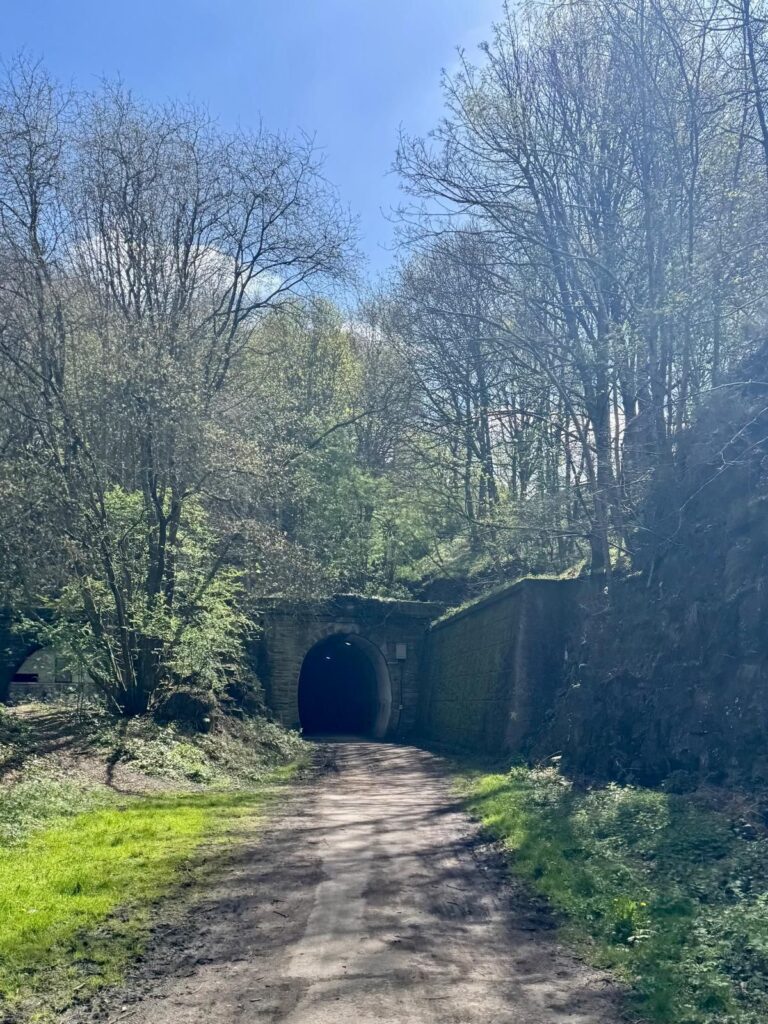 A sunny green and pleasant track heading towards a dark stone bridge and tunnel that looks almost black in contrast to its bright surroundings.