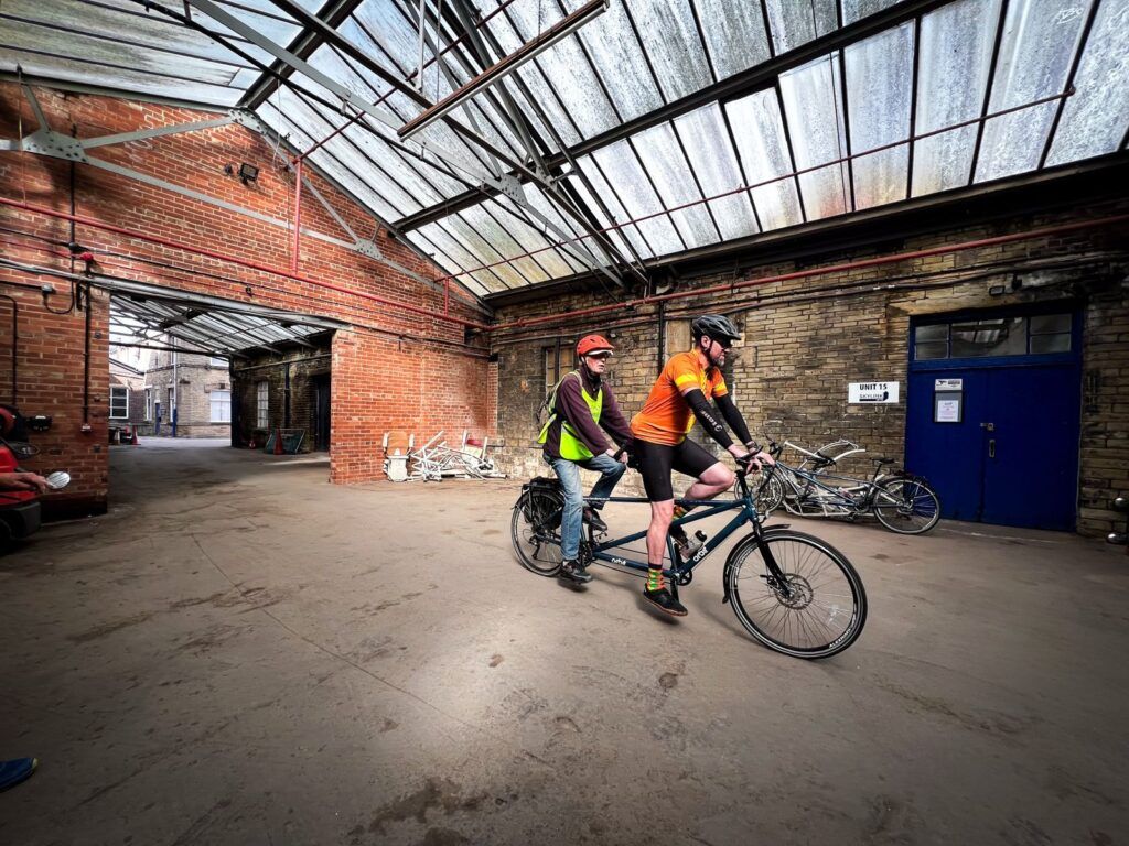 In a glass roofed industrial yard area, a pair ride on their tandem.