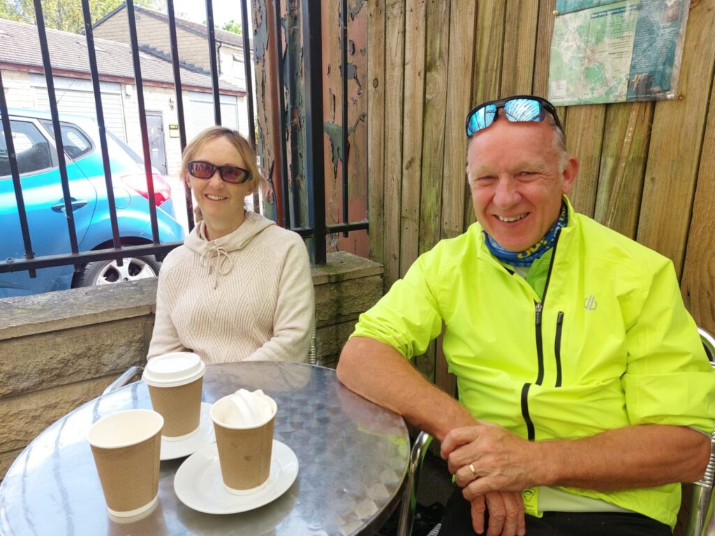  A man and woman sit at a metal bistro table smiling towards the camera, coffee cups are atop the table.
