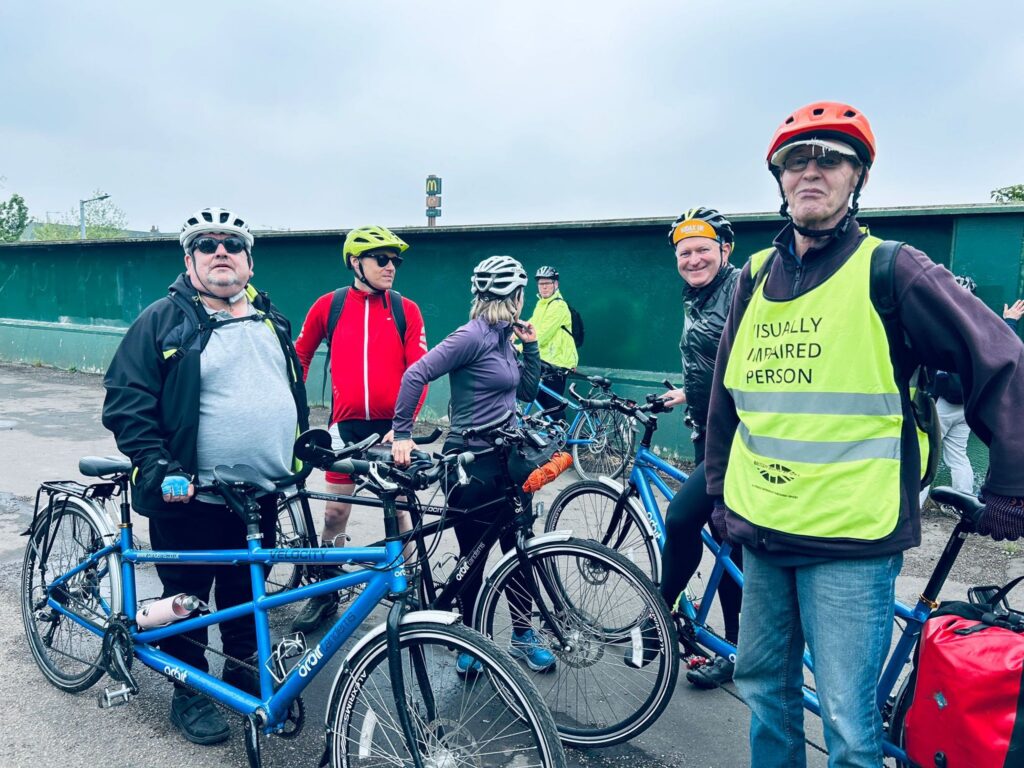 A group ftrekersstoodby ter bikes n a bridge with high solid green fences behind them.