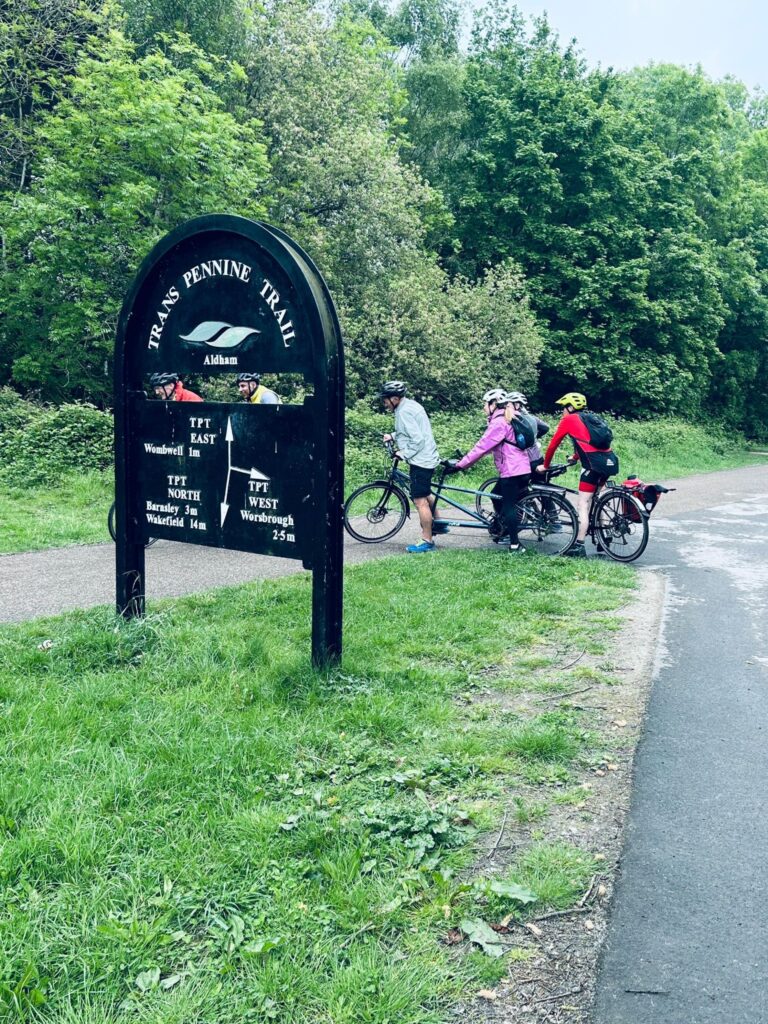 A fork in the path with several tandems taking the tight left turn going behind a big metal sign stating te Trans Pennine Trail