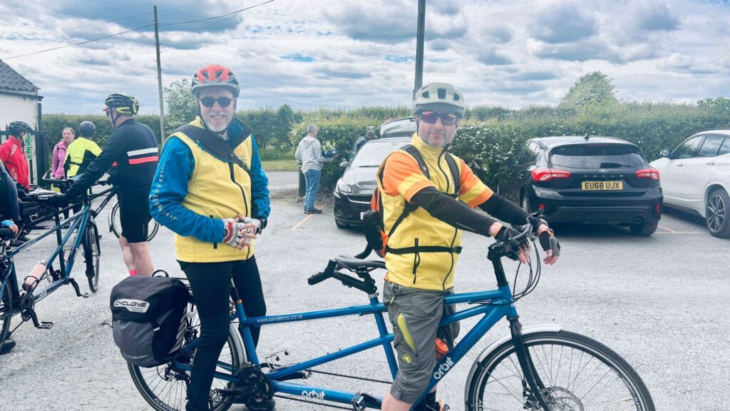 Two men are astride their blue tandem bicycle smiling in a sunny setting that appears to be a carpark.