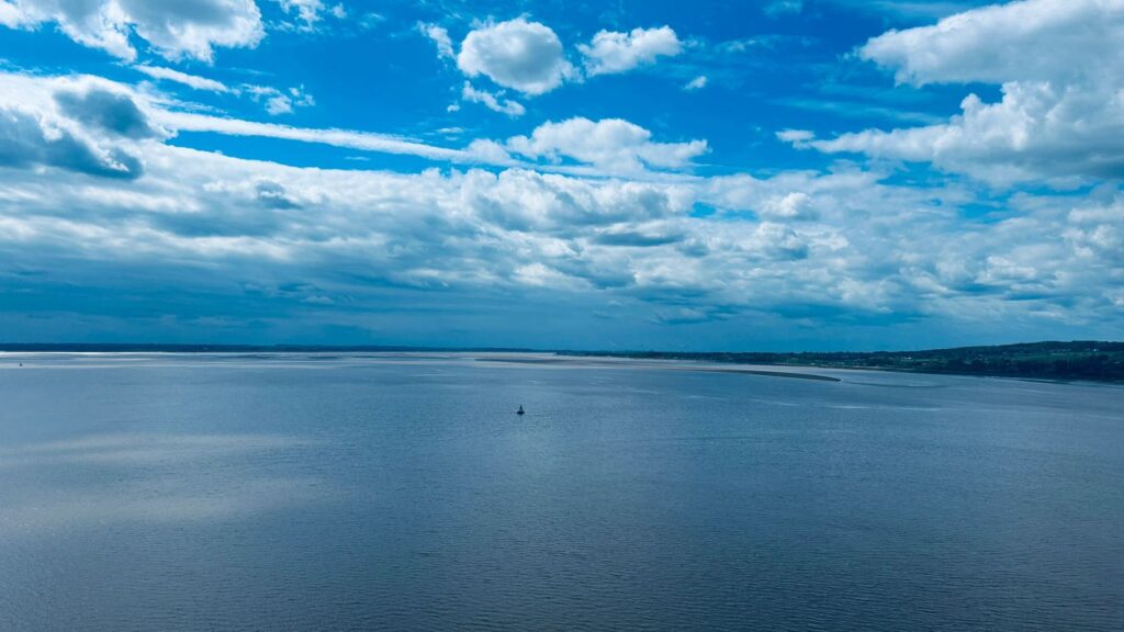 A sunny vista encompassing the wide Humber River glinting in the sunshine, with far off trees and foliage on the banks at either side. The sky is bue with fluffy white clouds. 