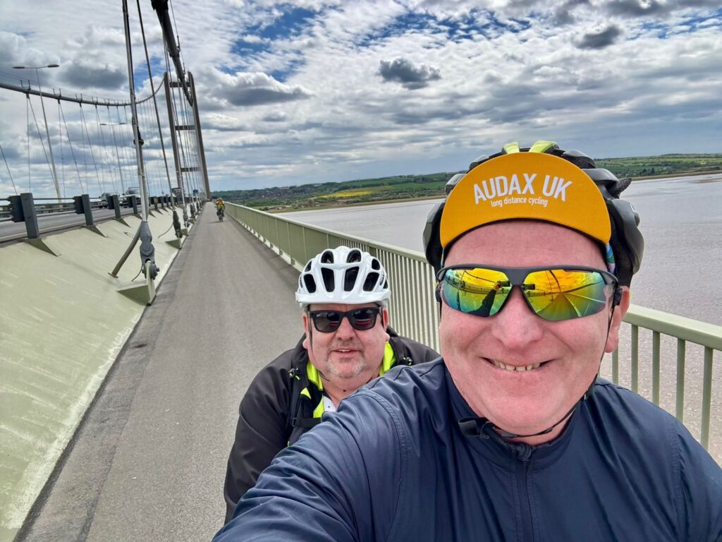 Steve and Martin in a selfie as they ride over the Humber Bridge. You can see the grey- brown water below with the iconic metal and concrete frame of the bridge visible in part behind them