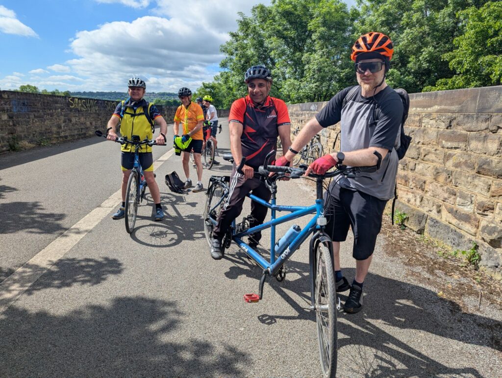 Several members of TT getting ready to get going. One pair are stood next to their tandem, another is arranging gear they have into a bag on the floor.