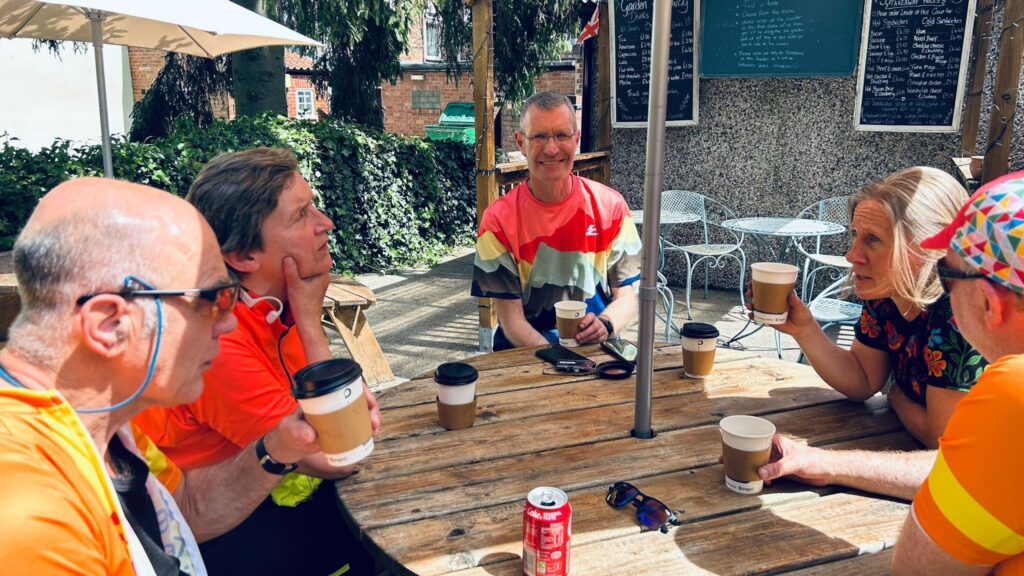 5 Trekkers, predominantly wearing hues of orange sit around a round wooden picnic table with a parasol. Evidence of refreshments are visible of the table.