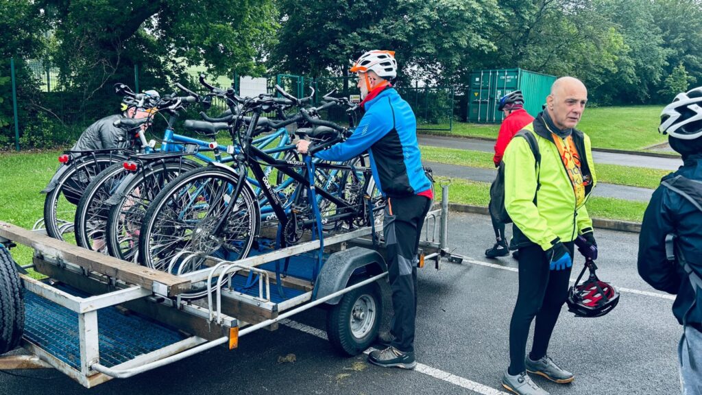 in a carpark area a cage like trailer is full of tandem bicycles. A tall man is lifting the nearest bike in (or out) of the trailer.