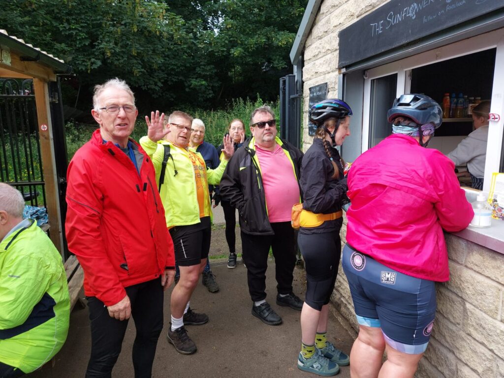 A group of Tandem Trekkers in various brightly coloured outfits queue up outside the cafe serving window. One member is leaning back stood up giving a double handed wave.