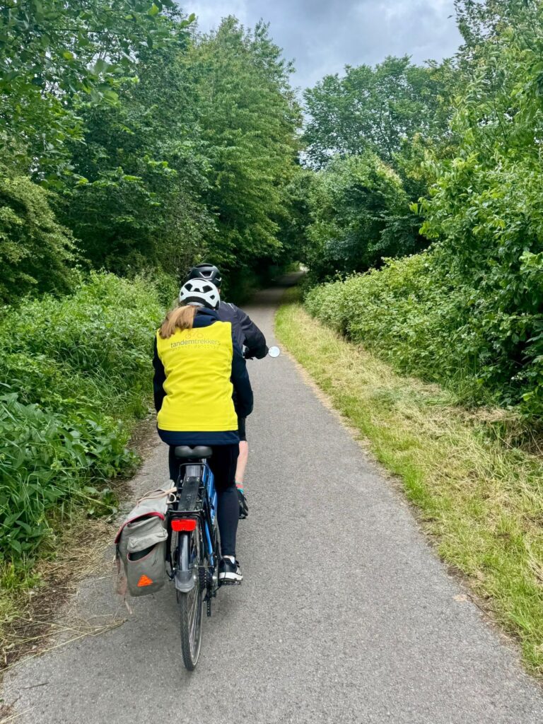 A tarmac path covered by a lush green tree canopy with a tandem riding away from the camera. The back rider has a yellow TT branded gilet on and the bike has a grey pannier on the left side by the back wheel.