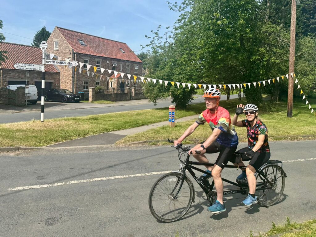 A tandem passes a small village green with stone buildings adorned in bunting. Stoker Lou is waving to camera smiling.