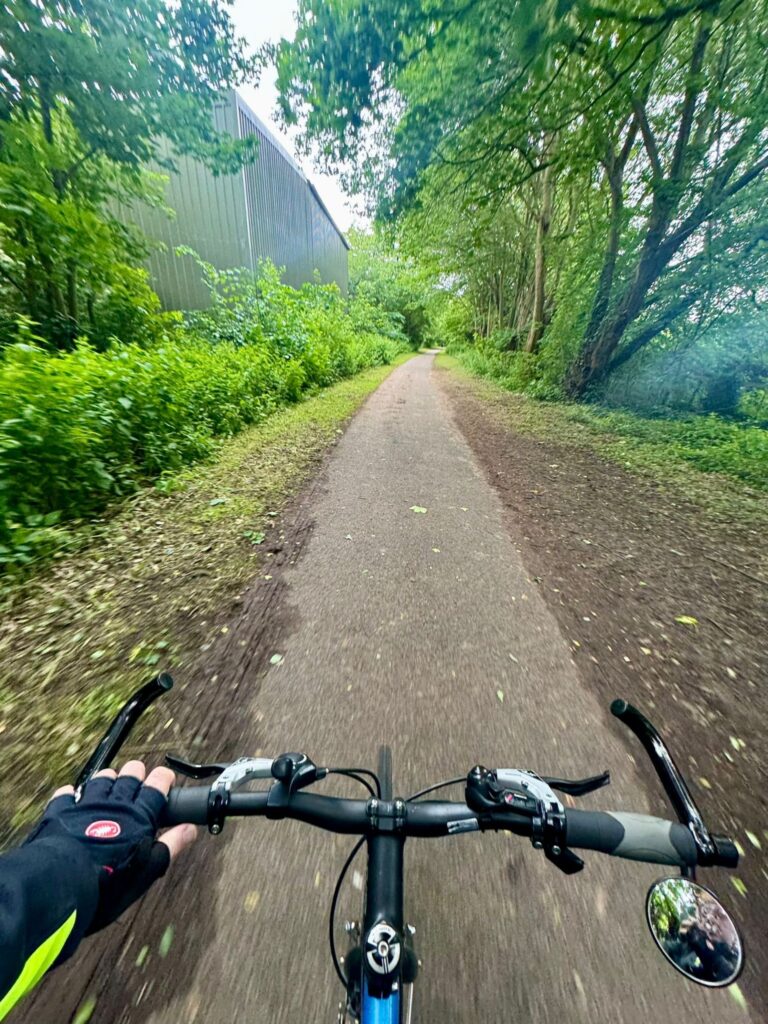 A p.o.v shot from the pilots position looking over the handle bars, a metal shed is to the left of a tarmac path with leaft green trees and dirt verges to the other side ad ging forward.