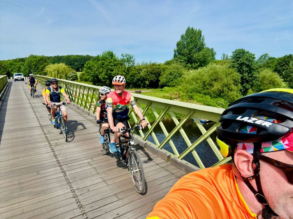 4 tandems are going across a straight wooden bridge with boards on the path and diamond patterned railings as the cross a river surrounded by large green trees