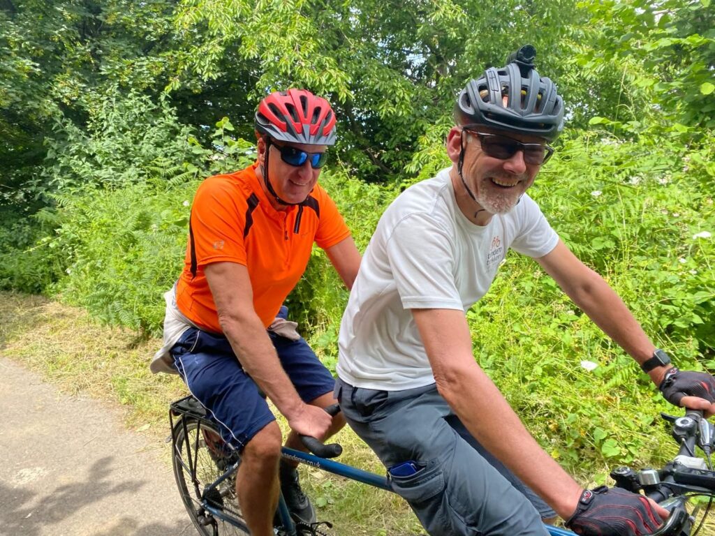 A thick green leafy backdrop with a pair on a tandem. The front pilot is approximately a 65 year old white male wearing a white tshirt. The stoker is of similar age and ethnicity but wearing orange.