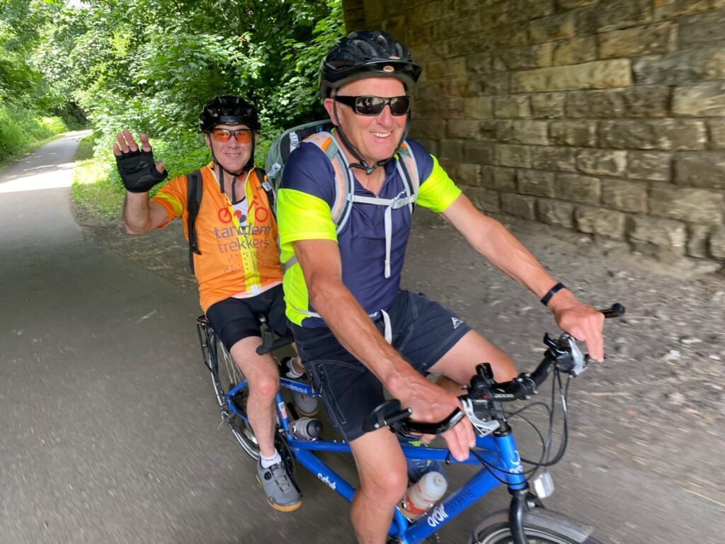 Background shows a stone bridge wal which the cyclists are going under. There is one tandem with a pair of smiling riders. The stoker is waving at the camera with their right hand.