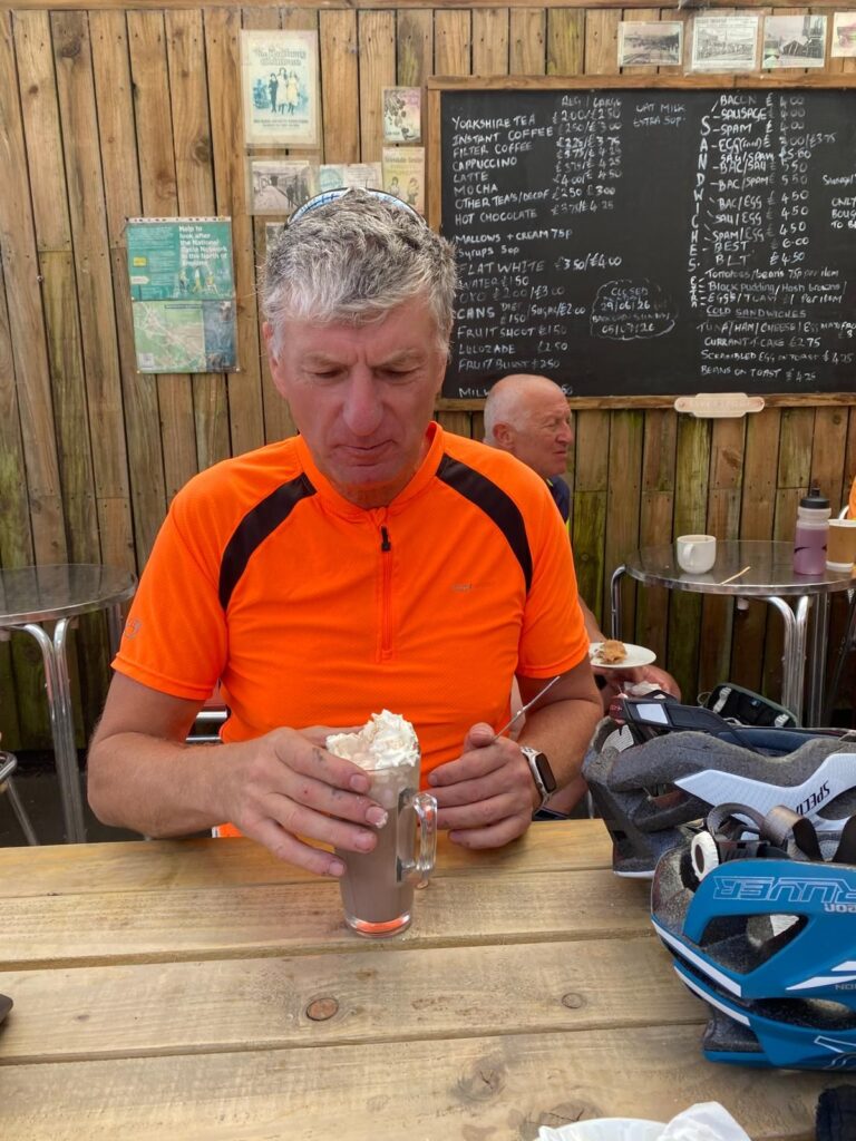 A man in an orange cycle top sits at a wooden table with a large hotchocolate drink infront of him. It is topped with a generous quantity of cream. His helmet sits to the side of him atop the table.