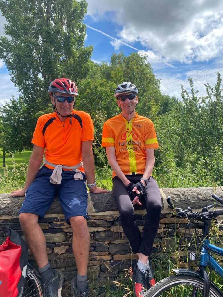 Two men in orange cycling tops sit atop a stone wall in bright sunshine laughing. Parts of their bies which are laid on the floor infront of the low wall are visible. They are surrounded by tall grassed and vergeside plants.