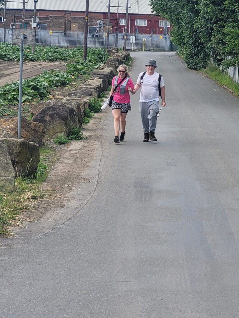 A long straight stretch of road shows two people waking side by side, they have shorts, shirts and hats on. There are low level plans to the left of the picture separated from the road by big sandstone boulders.