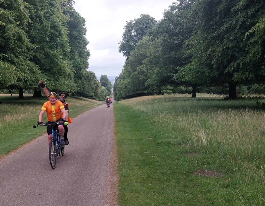 A tandem draws close with two smiling riders on it, the view shows a long straight path that goes off to the horizon, surrounded by grassy parkland and mature trees in verdant green leaf. More tandems can be seen in the distnce at intervals. The whole stretch of path show what is probably a half mile distance.