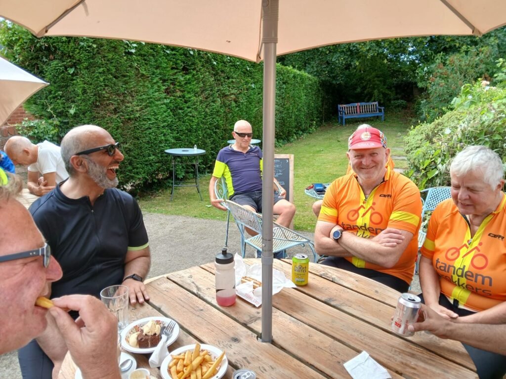 A garden style setting, 5 trekkers sit around a wooden picnic yable with parasol ejoying food ad drink. Sveral members have big grins or are laughing ope mouthed.