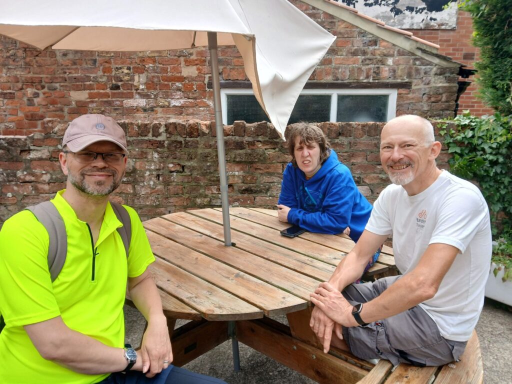 Steve G and Stevie B sit at a round wooden picnic table half tuned towards the camera smilong and posing. Katie is behind the table leaning her elbows across the top to join the photo. 