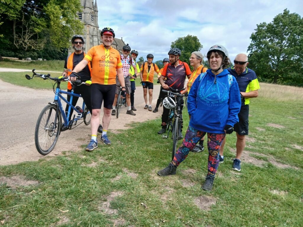 A group shot with some members still on bikes while others stand on their own two legs. They are clustered together in a grassy parkland area with mature trees and an ornate building with a stone spire in the background.