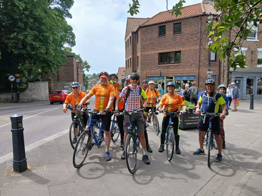 In a town street setting on a wide pavement rea withstreet furniture and planting the group pose for a photo. They are all stood but in position on their tandem bicycles.
