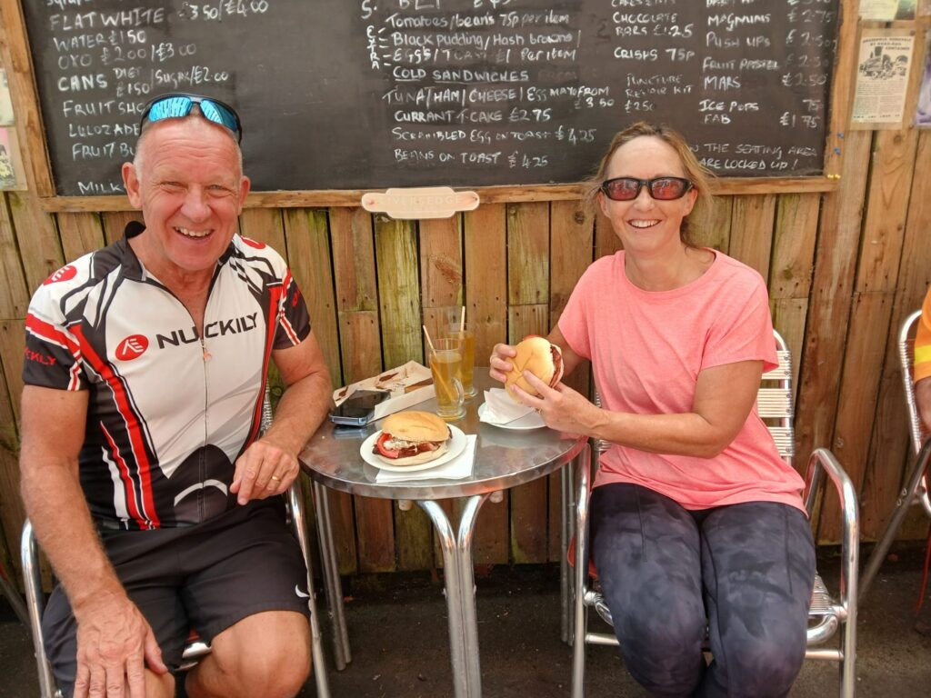 A man and a woman sit either side of a chrome bistro table. They are both smiling at the camera. The woman is poised to eat her sandwich.
