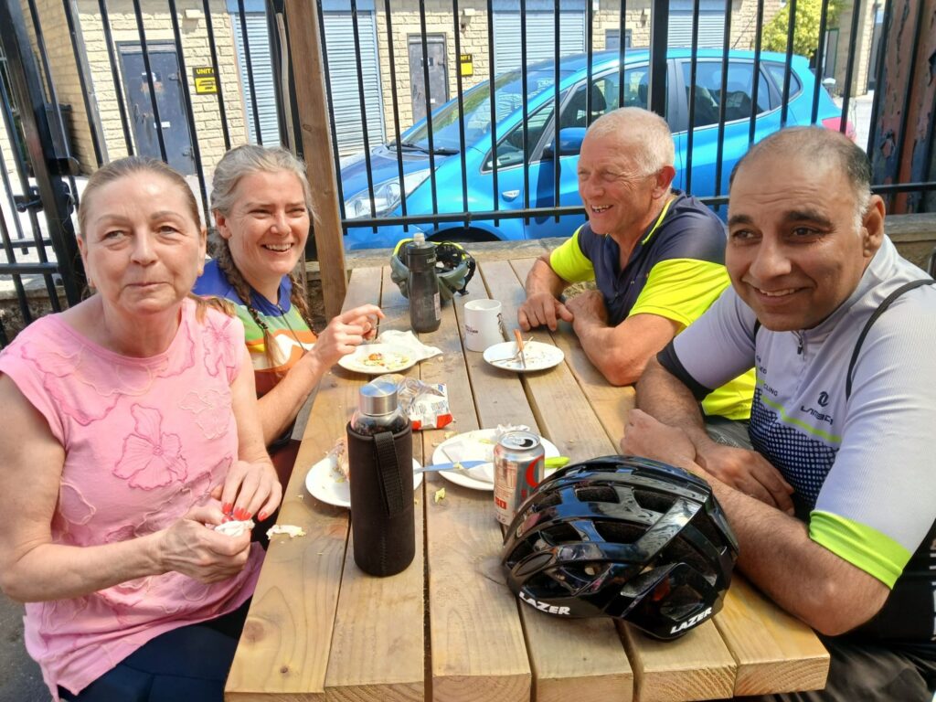 A group of 4, 2 male ad 2 female, sit opposite each other on a wooden picnic table. There are cycling helmets, water bottles and empty plates in front of them.