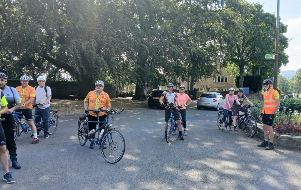 4 tandems are visible with their riders still mounted, paused haphazardly in a dappled shaded area. The ride leader, a man without bike, in TT hi-vis looks on ensuring everyone is present and safe 