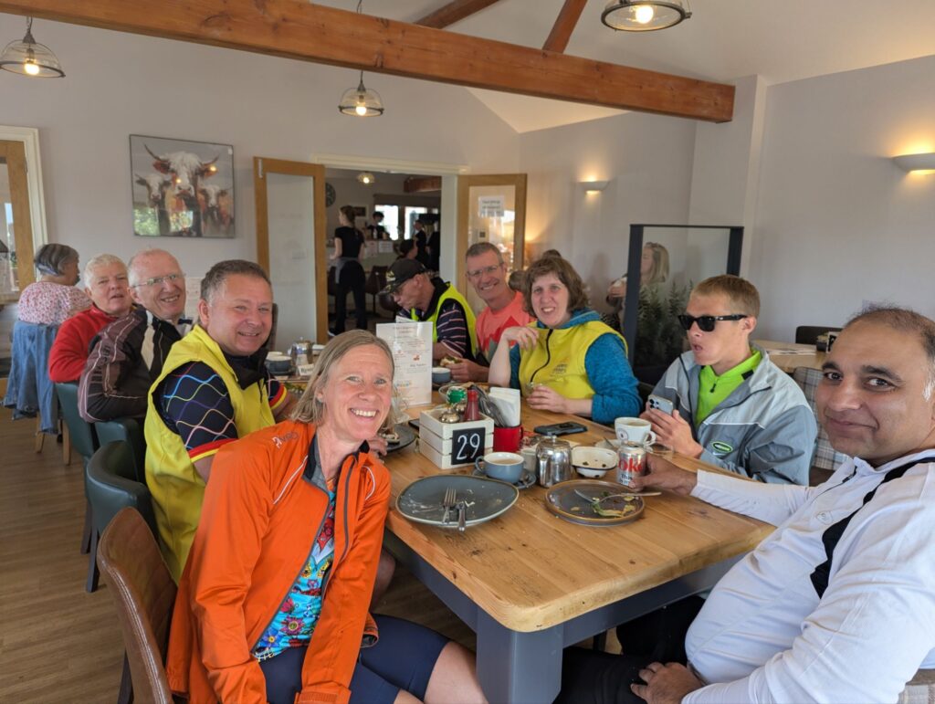 A group of trekkers sit around a large farmhouse style table in the centre of a busy cafe. The room is light and airy with white walls and exposed pine coloured beams. The group are all sat looking at the camera smiling with empty plates and cups infront of them onthe table.